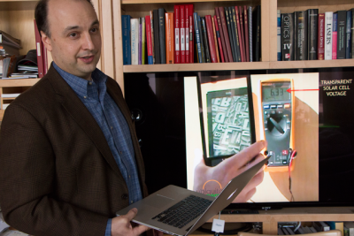 Professor Vladimir Bulović discusses research that led to transparent solar cells, which can power an electronic book reader, as shown in video on television screen behind him. An MIT spinoff, Ubiquitous Energy, based in Redwood City, California, is commercializing the technology. Photo: Denis Paiste/Materials Processing Center